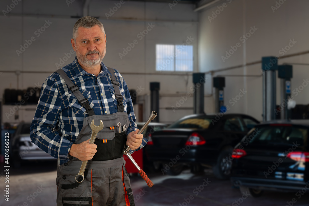Portrait of an elderly auto mechanic. The experienced man with a beard is wearing work trousers and has a wrench in his hands. In the background are cars and lifting platforms. Portrait of an elderly auto mechanic. The experienced man with a beard is wearing work trousers and has a wrench in his hands. In the background are cars and lifting platforms.