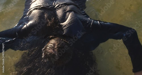 Drowned man with long dark hair in the water sways in the muddy water lying face down in shallow water. Unrecognizable body in black clothes is washed ashore after a shipwreck. 50 fps 4k slow motion