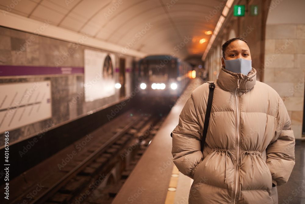 Portrait of young woman in protective mask looking at camera while standing in underground with train in the background Portrait of young woman in protective mask looking at camera while standing in underground with train in the background