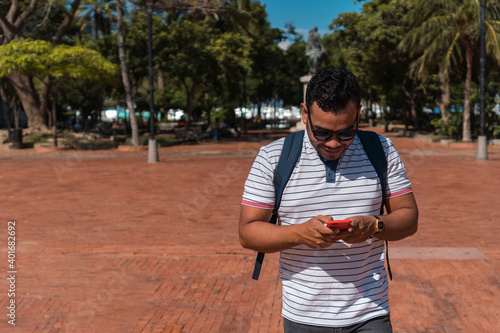 Young Latino looking at his cell phone and smiling - Young Hispanic texting online at the outdoor park - New technology trends addiction concept