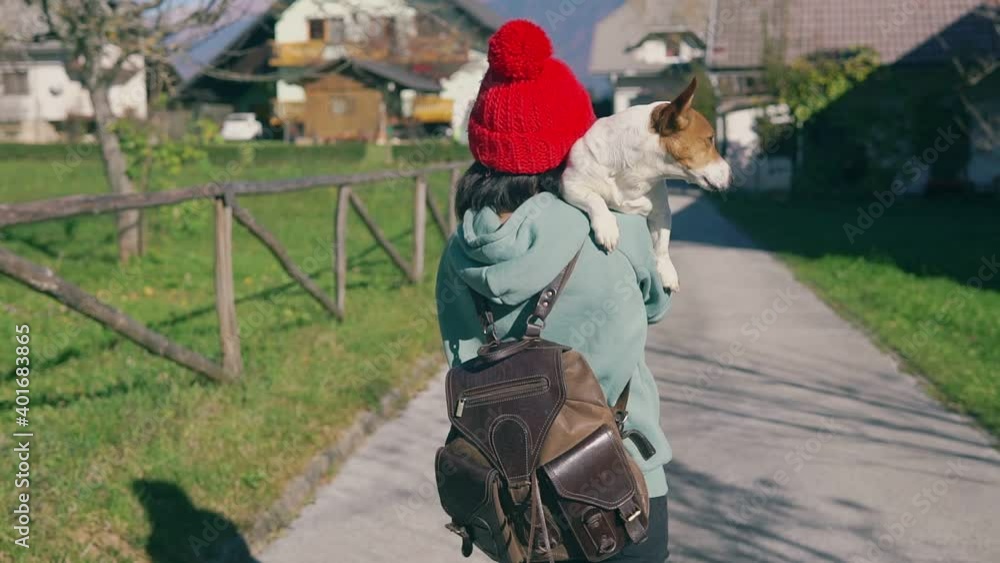 Happy owner and her doggy spend time together. Young girl in red cap carrying a small dog on her shoulder and walking at countryside on grass lawn. Concept of domestic animals and pets Happy owner and her doggy spend time together. Young girl in red cap carrying a small dog on her shoulder and walking at countryside on grass lawn. Concept of domestic animals and pets