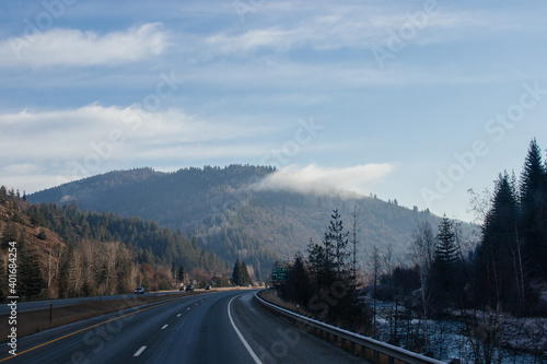 An asphalt road along which cars and trucks go, among the mountains, ahead on the horizon mountains in fog, the sky is covered with clouds. Idaho, USA, 11-23-2019