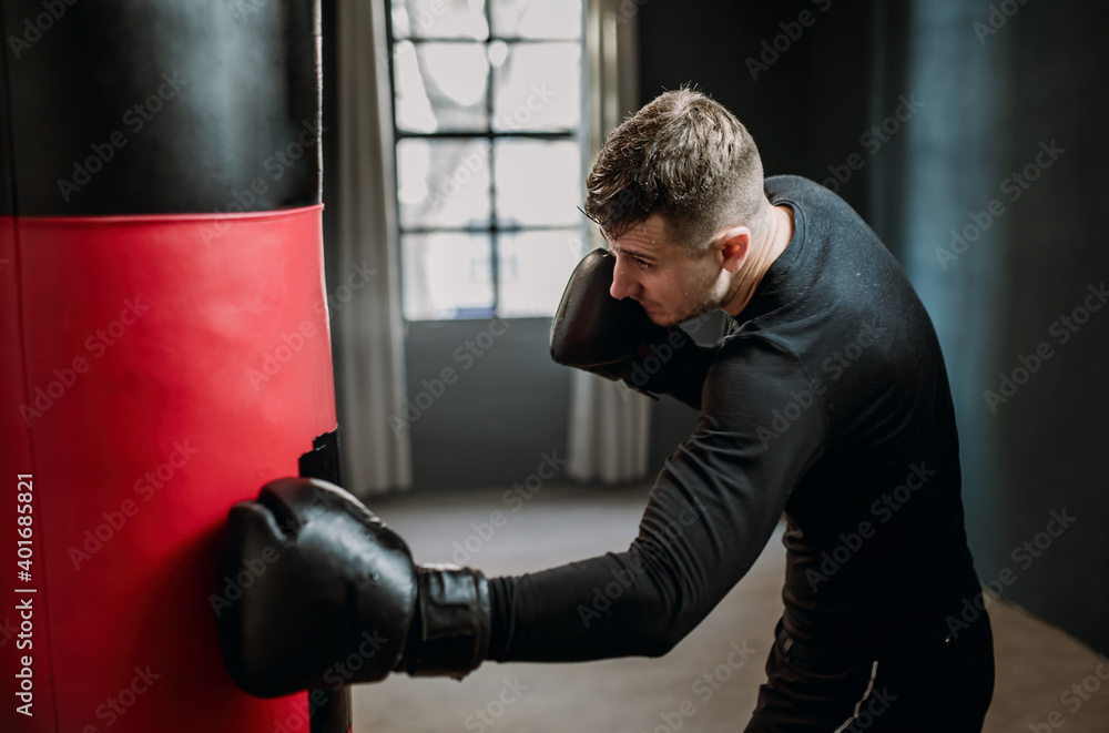 Boxing, athletic man fighter trains his punches, beats a punching bag, training day in the boxing gym, strikes with his feet, back lighting.