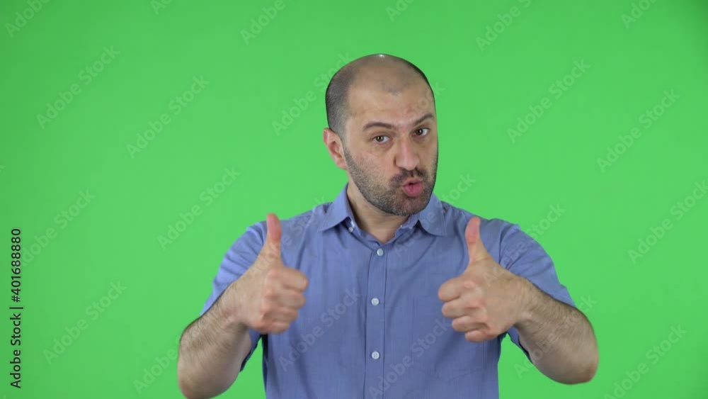 Portrait of a middle aged men looking at the camera shows a thumbs up gesture with both hands. Balding male with beard in blue shirt posing on green screen in the studio. Close up. Portrait of a middle aged men looking at the camera shows a thumbs up gesture with both hands. Balding male with beard in blue shirt posing on green screen in the studio. Close up.