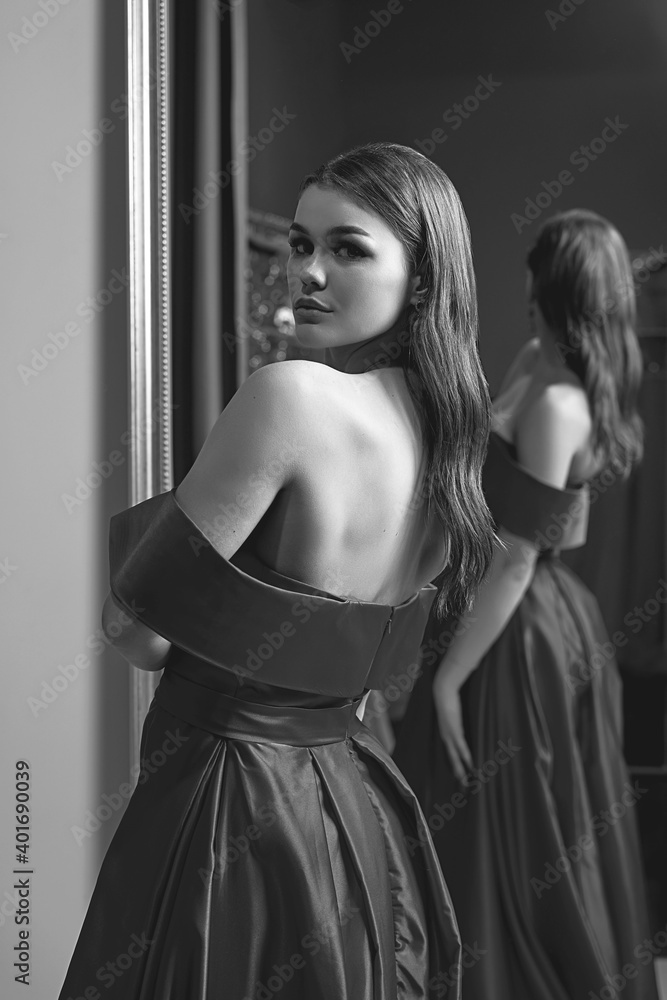 Young beautiful girl wearing an off-the-shoulder full-length satin slit prom ball gown. Model looking in mirror. Black and white image in retro style.