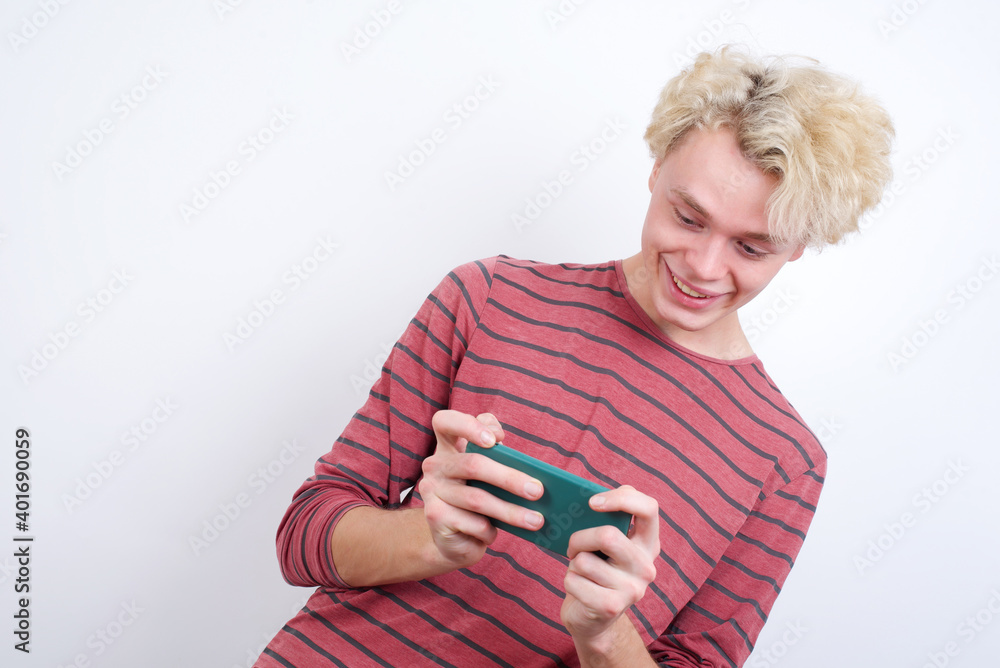 Young handsome Caucasian blond man standing against white background holding in hands cell playing video games or chatting Young handsome Caucasian blond man standing against white background holding in hands cell playing video games or chatting