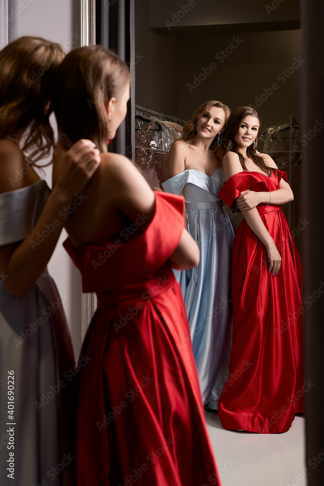 Two young beautiful girls wearing off-the-shoulder full-length sky blue and crimson red satin slit prom ball gowns. Models looking in mirror. Fitting room in dress hire service.