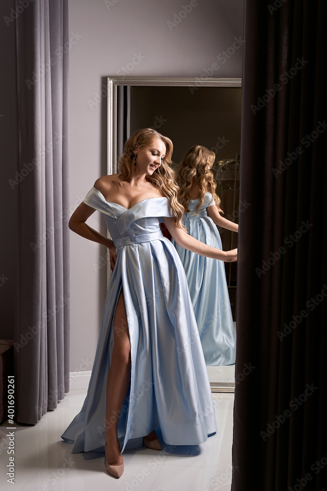 Young beautiful blonde girl wearing an off-the-shoulder full-length sky blue satin slit prom ball gown. Model looking in mirror. Fitting room in dress hire service.