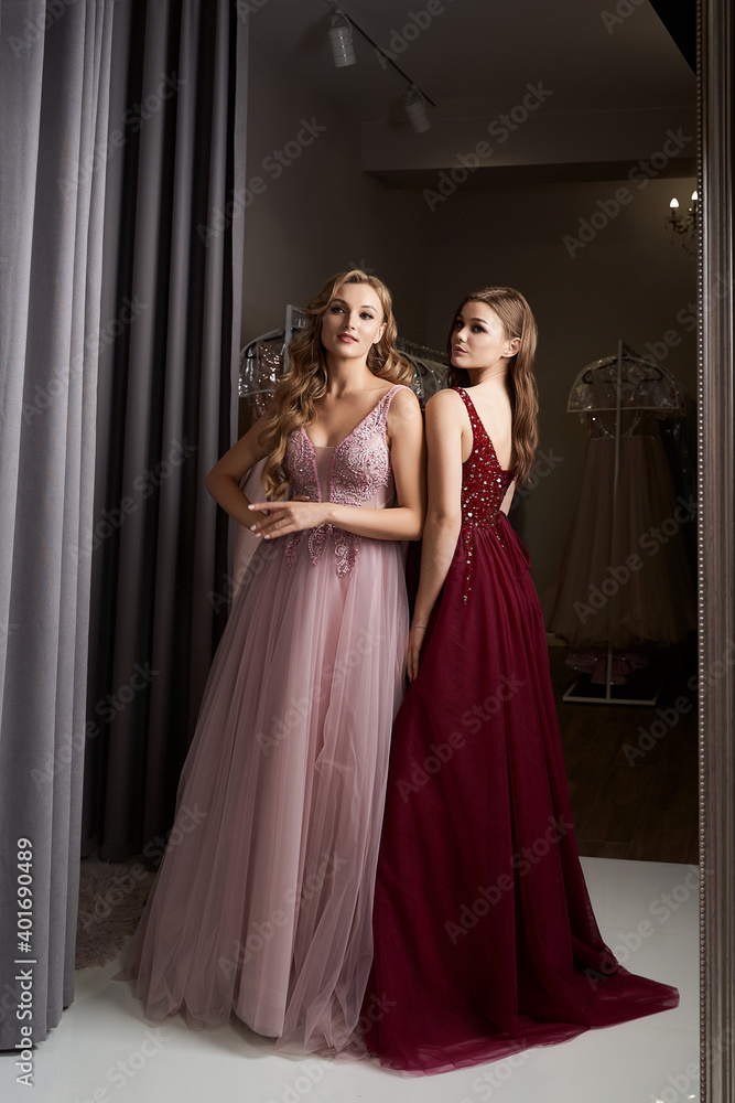 Two young beautiful girls wearing a full-length dark crimson red chiffon prom ball gown decorated with sparkles and sequins. Model in front of mirror in a fitting room at dress hire service.