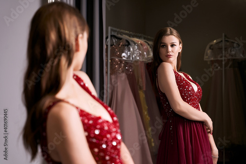 Young beautiful brunette girl wearing a full-length dark crimson red chiffon prom ball gown decorated with sparkles and sequins. Model in front of mirror in a fitting room at dress hire service.
