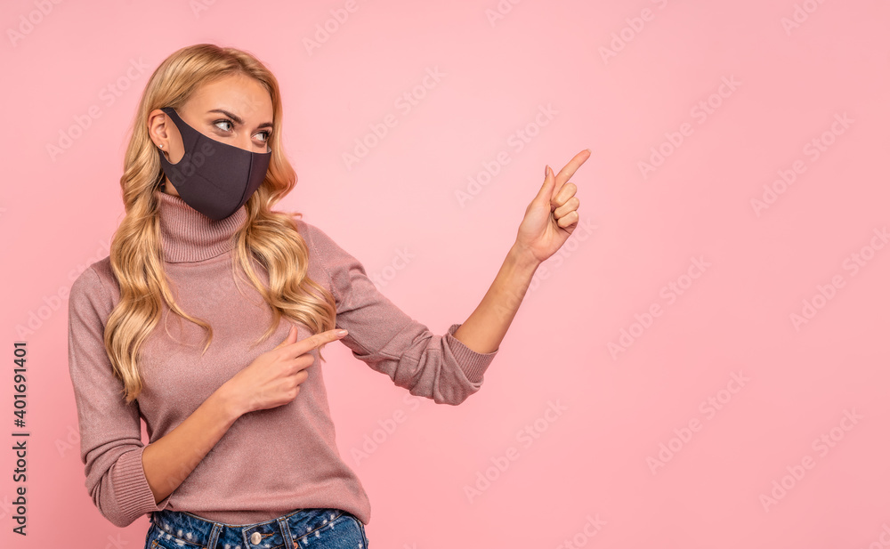 Young woman in pink blouse, sterile face mask to safe from coronavirus  virus covid-19 during pandemic quarantine pointing fingers on workspace  mock ups copy space isolated on pastel pink background. Stock Photo