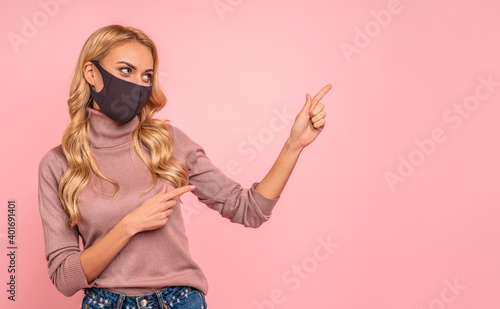 Young woman in pink blouse, sterile face mask to safe from coronavirus virus covid-19 during pandemic quarantine pointing fingers on workspace mock ups copy space isolated on pastel pink background.