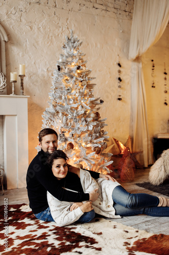 Adorable couple lying down near decorated Christmas tree in bedroom, smiling. Loving husband hugging beautiful wife, enjoy tenderness. Handsome man celebrate xmas eve with gorgeous woman at home