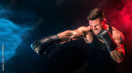 Bearded tattooed sportsman muay thai boxer in black undershirt and boxing gloves fighting on dark background with smoke. Sport concept. © zamuruev