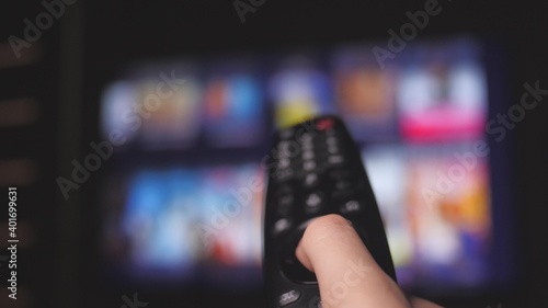 Woman hand selects internet tv channels with remote control, close-up. Person controls TV using a modern remote control. Girl watches smart TV and uses black remote control. Blurry tv scrolls pages