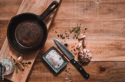 Iron pan, on a kitchen table, accompanied by seasonings, salt with spices, kitchen knife, garlic, and fresh oregano on a rustic madara background