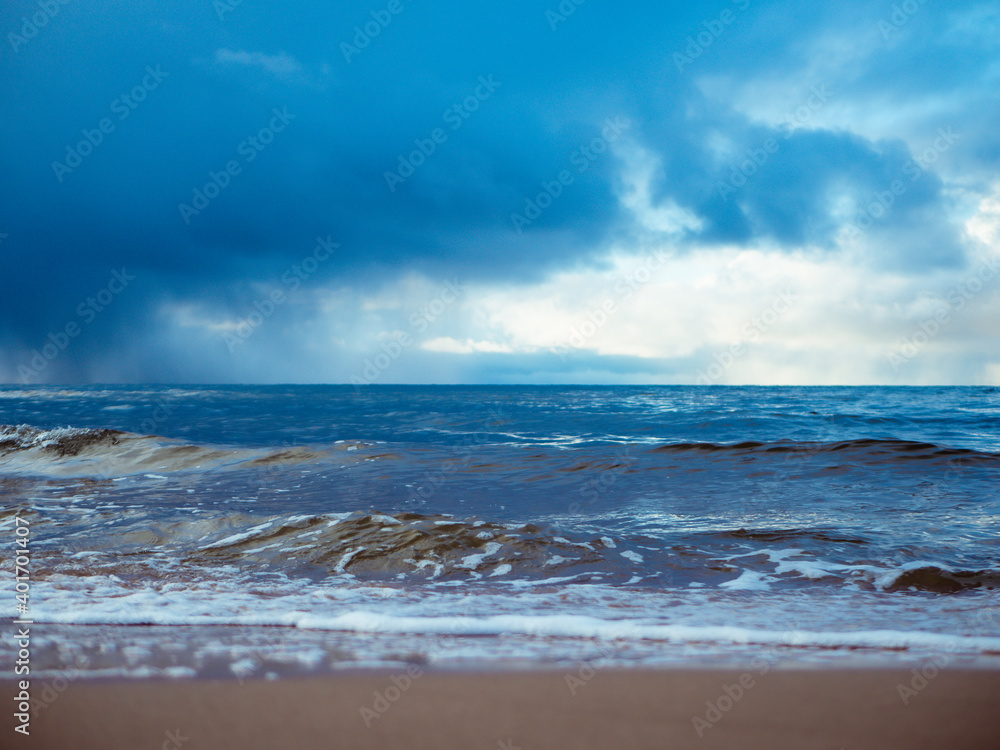 The small waves of the sea fight with the sand. Sea waves wash out the sandy shore. Rain clouds are visible in the sky