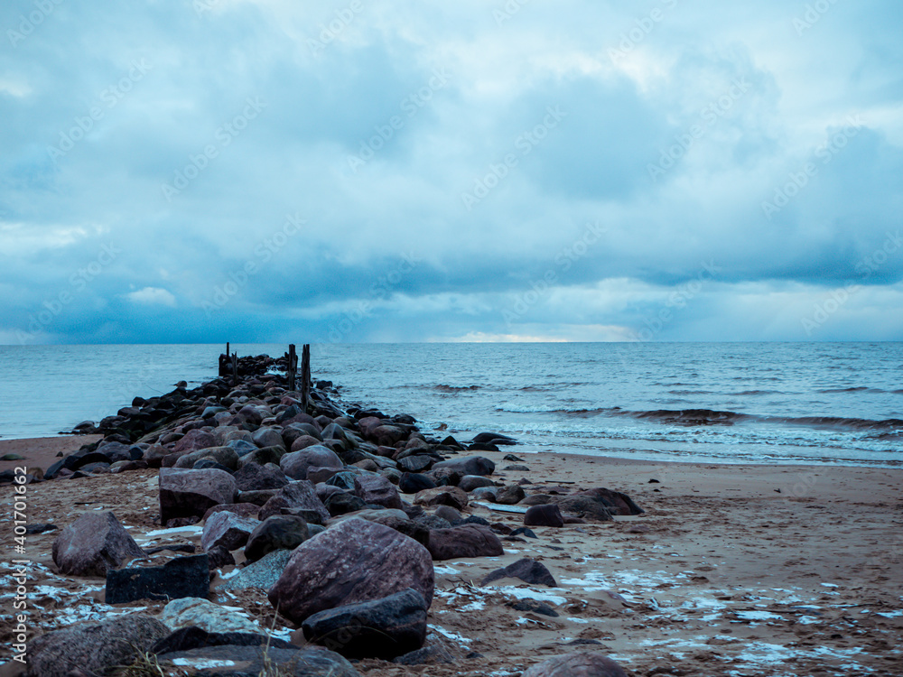 The sea wave washes away the old abandoned stone pier. Rain clouds are visible in the sky.