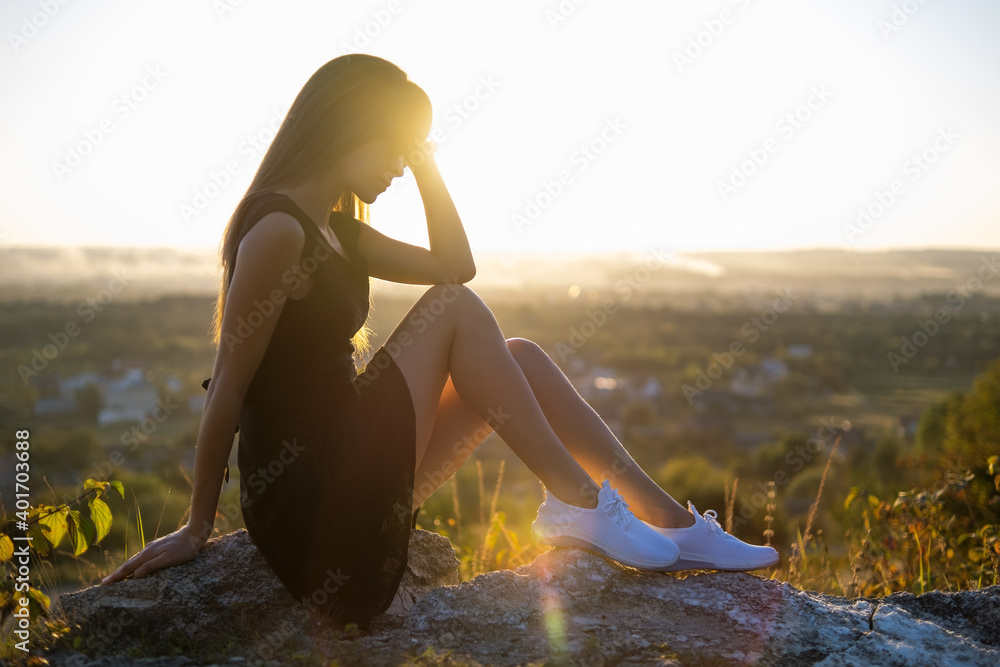 Young elegant woman in black short dress and white sneaker shoes sitting on a rock relaxing outdoors at summer evening. Fashionable lady enjoying warm sunset in nature. - obrazy, fototapety, plakaty 
