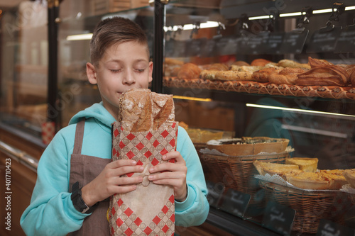 Lovely young boy wearing apron, smelling fresh bread at his family bakery shop