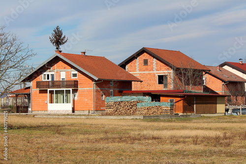 Row of newly built unfinished red brick family houses with new roof tiles surrounded with dry grass and small wooden outdoor storage sheds on cloudy cold winter day