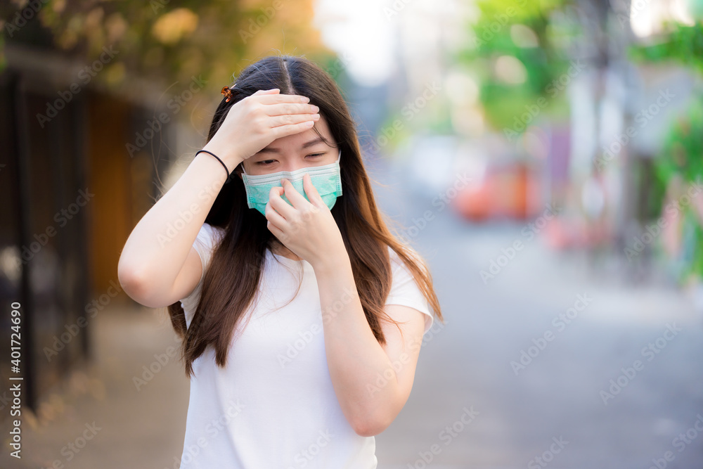 Teenage woman in a green medical face mask is coughing, and she touches her forehead with her hands to take body temperature. Prevent the spread of germs.