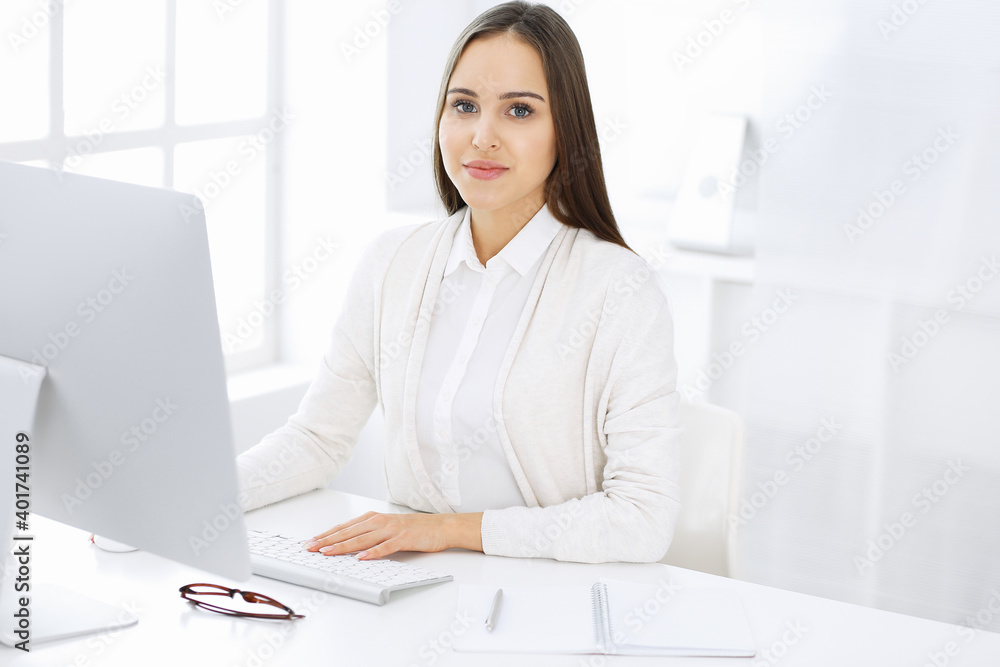 Young cheerful woman sitting at the desk with computer and looking at camera in white colored office. Looks like student girl or business lady. Headshot Young cheerful woman sitting at the desk with computer and looking at camera in white colored office. Looks like student girl or business lady. Headshot