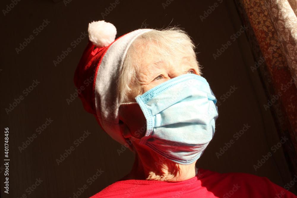 Fototapeta premium Senior woman near the window in the sunny morning with a Santa hat wears a medical mask due to the coronavirus.