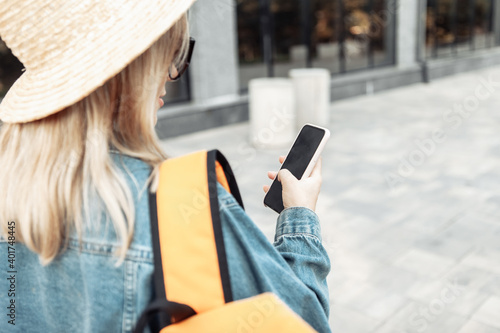Young millennial woman tourist in sunglasses and hat with tourist backpack uses smartphone in city