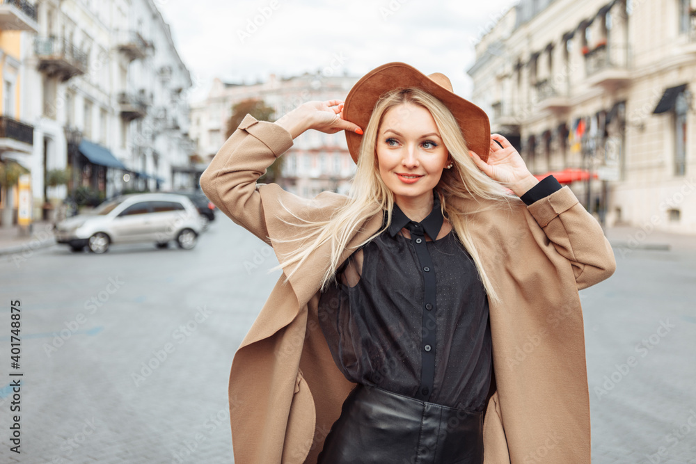 Portrait of young stylish blonde woman in autumn coat and felt hat in a European city. Beautiful attractive girl