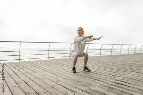 Cute blonde sportswoman trains on the beach. Young slim fit girl is practicing squats. Healthy lifestyle, fitness concept