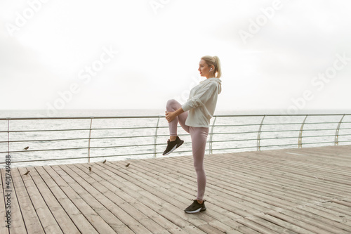 Fototapeta Naklejka Na Ścianę i Meble -  Fit blonde woman practicing leg stretching before outdoor workout. Young girl dressed in white hoodie trains on beach. Healthy lifestyle, sports and fitness
