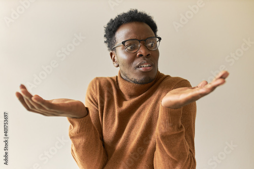 Frustrated young African man in trendy glasses shrugging shoulders in bewilderment, having confused doubtful look, spreading palms, feeling uncertain about making decision, weighing all pros and cons photo