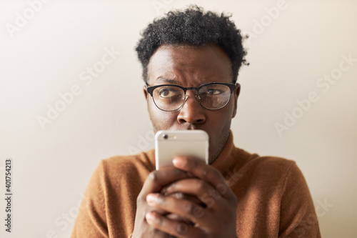 Electronic gadgets and online communication. Studio image of suspicious young dark skinned man typing text message via online messenger on mobile. Thoughtful black guy reading sms, texting reply photo