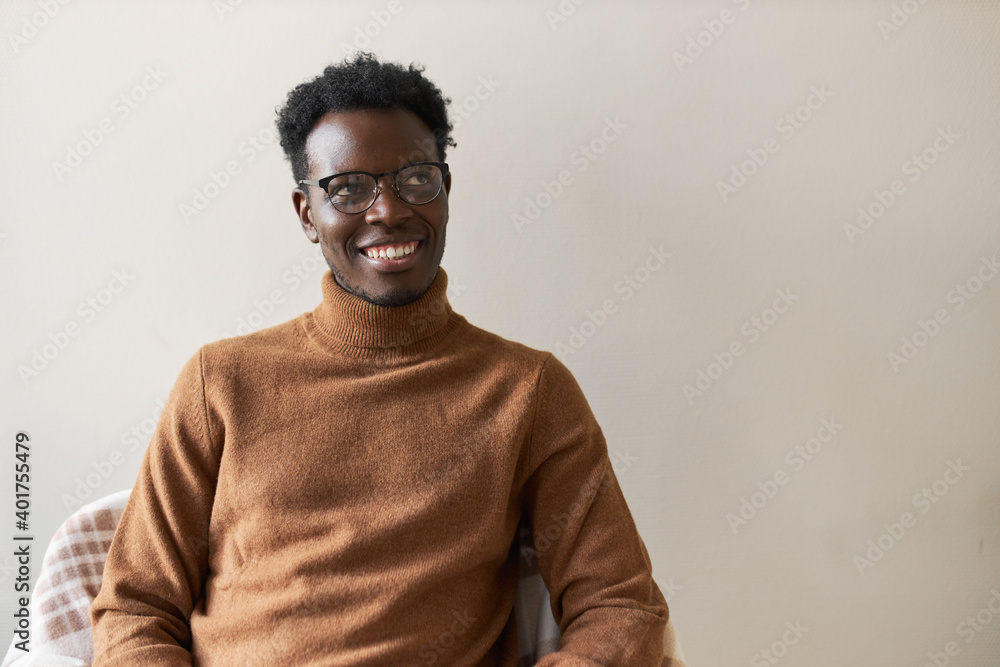Obraz premium Attractive cheerful young dark skinned unshaven man sitting on chair, waiting for his turn in queue for job interview, smiling joyfully, feeling confident, wearing casual sweater and spectacles