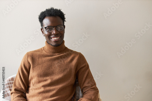 Joy, positive emotions and happiness concept. Enthusiastic successful young black businessman in eyewear and cashmere sweater looking away with happy joyful smile, being proud of himself © Anatoliy Karlyuk