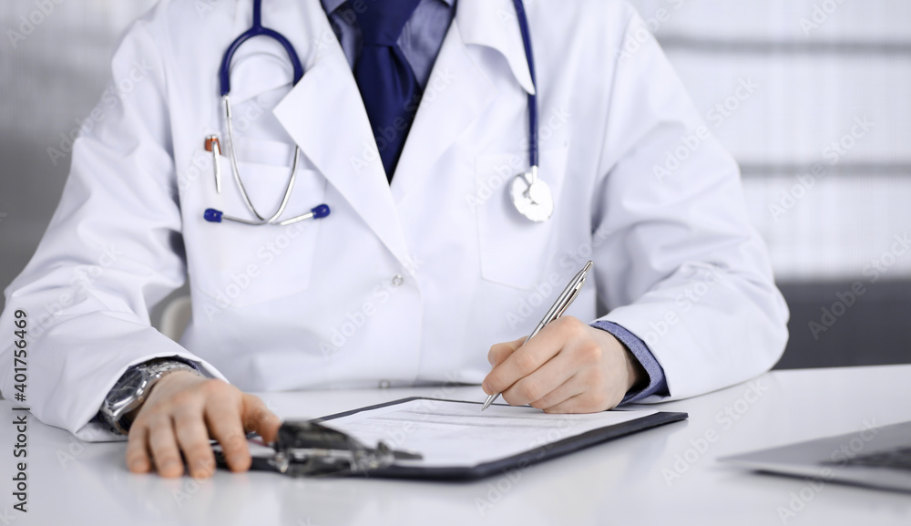 Unknown male doctor sitting and working with clipboard in clinic at his working place, close-up. Young physician at work. Perfect medical service, medicine concept Unknown male doctor sitting and working with clipboard in clinic at his working place, close-up. Young physician at work. Perfect medical service, medicine concept
