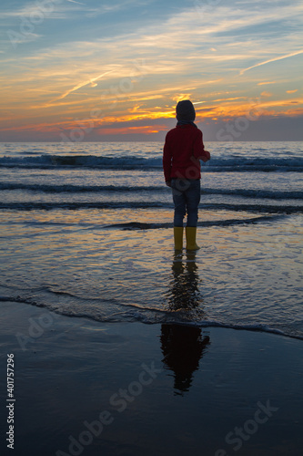 silhouette of a xoung boy standing at the shore of the north sea in the netherlands with waves breaking and a beautiful sunset in the background