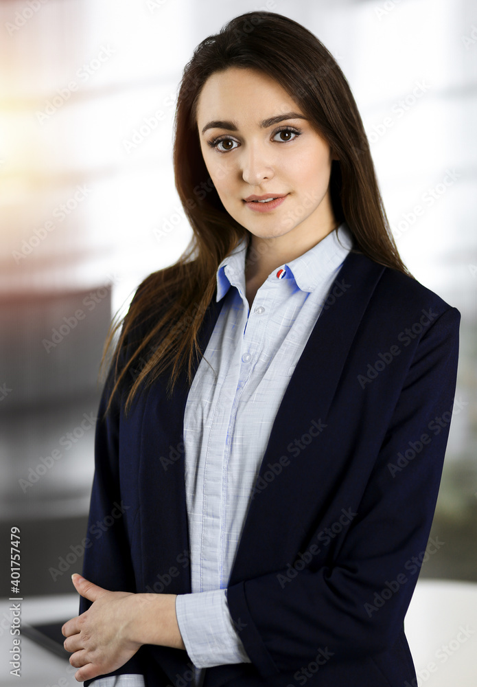 Intelligent young business woman in a blue jacket is standing at her workplace. Portrait of a specialist in sunny office Intelligent young business woman in a blue jacket is standing at her workplace. Portrait of a specialist in sunny office
