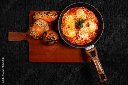 Shakshuka in a Frying Pan. Eggs Poached in Spicy Tomato Pepper Sauce. Homemade breakfast shakshuka - fried eggs, onion, bell pepper, tomatoes and parsley in a pan on a rustic black table  photo