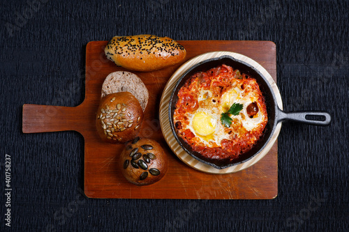 Shakshuka in a Frying Pan. Eggs Poached in Spicy Tomato Pepper Sauce. Homemade breakfast shakshuka - fried eggs, onion, bell pepper, tomatoes and parsley in a pan on a rustic black table  photo