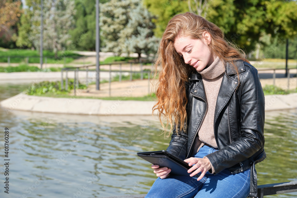 Redhead caucasian woman reading or working in a e-book or tablet in a park. Digital nomad entrepreneur concept. Redhead caucasian woman reading or working in a e-book or tablet in a park. Digital nomad entrepreneur concept.