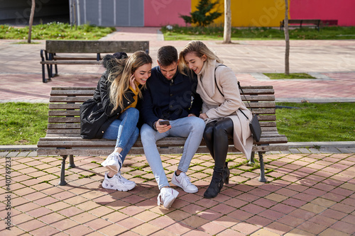 Three young friends sitting outdoors and looking at their cell phones. A group of people sitting on a park bench outdoors and watching a video on their smartphone. Concept of friendship and technology