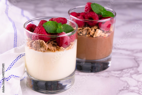 Two glasses of vanilla and chocolate pudding, garnished with raspberries and mint on a marble table.