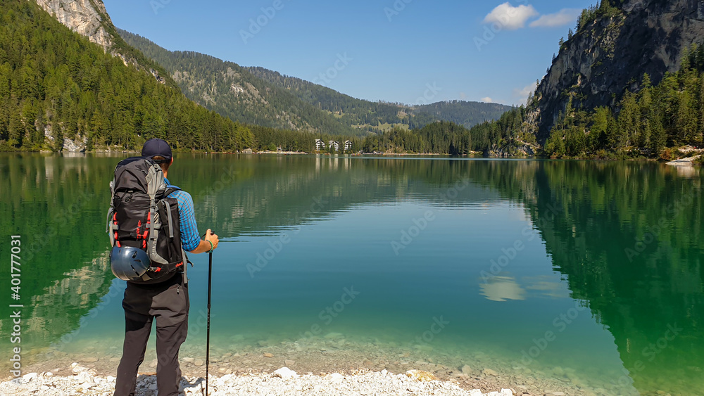 A man with a big backpack standing at the shore of the Pragser Wildsee, a lake in South Tyrolean Dolomites. High mountain chains around the lake. The sky and mountains are reflecting in the lake A man with a big backpack standing at the shore of the Pragser Wildsee, a lake in South Tyrolean Dolomites. High mountain chains around the lake. The sky and mountains are reflecting in the lake
