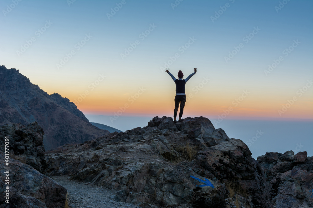 A female tourist standing on the peak of a rocky mountain watching sun rising in Darband valley in autumn in dawn against colorful sky in the Tochal mountain. A female tourist standing on the peak of a rocky mountain watching sun rising in Darband valley in autumn in dawn against colorful sky in the Tochal mountain.