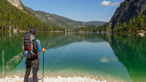 A man with a big backpack standing at the shore of the Pragser Wildsee, a lake in South Tyrolean Dolomites. High mountain chains around the lake. The sky and mountains are reflecting in the lake