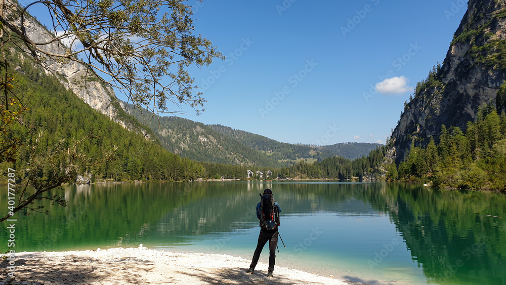 A man with a big backpack standing at the shore of the Pragser Wildsee, a lake in South Tyrolean Dolomites. High mountain chains around the lake. The sky and mountains are reflecting in the lake