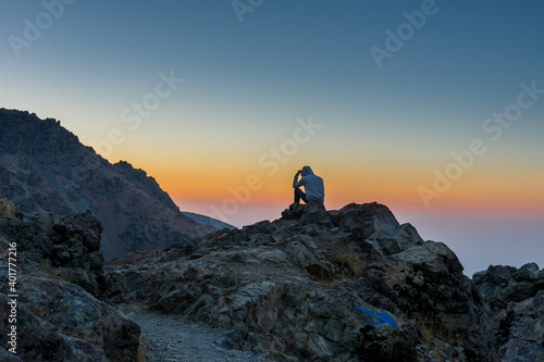 A tourist sitting on the peak of a rocky mountain watching sun rising in Darband valley in autumn in dawn against colorful sky in the Tochal mountain.