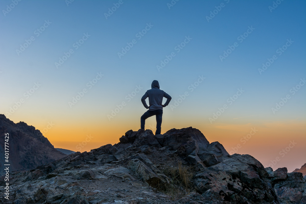 A male tourist standing on top of rocky mountain watching sun rising in Darband valley in autumn in dawn against colorful sky in the Tochal mountain.